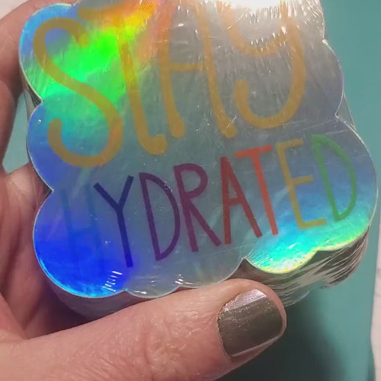 shiny shiny kiss-cut holographic sticker reads 'stay hydrated' in bold yellow and rainbow lettering on a reflective rainbow-silver holographic background